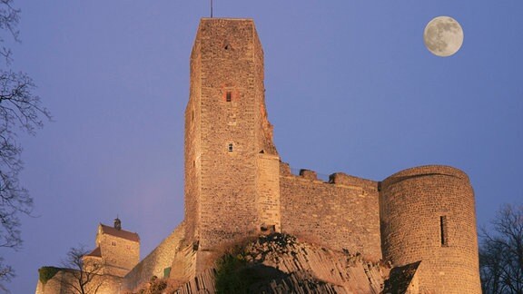 Burg Stolpen, eine Burganlage auf einem Felsen in der Dämmerung, am Himmel ist der Mond zu sehen.