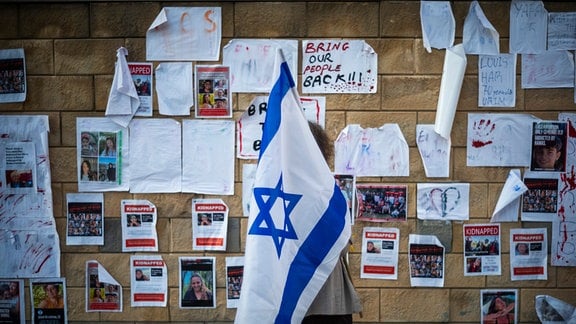 Eine Frau, die eine israelische Fahne hält, guckt sich Plakate an, auf denen Personen zu sehen sind, die von der Hamas entführt wurden.