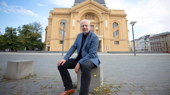 Neue Spielzeit am Theater Altenburg Gera: ein Mann sitzt vor einem Theatergebäude auf einem Stein.