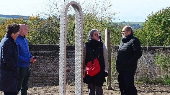 Einweihung des Kunstwerkes "Bogen" von Leunora Salihu für den Purple Path in Stollberg: Drei Menschen betrachten den schmalen, silberglänzenden Bogen. Im Hintergrund ist Landschaft zu sehen. 