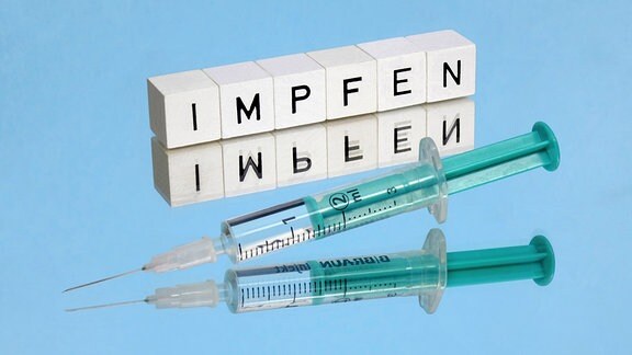 Buchstabenwürfel mit dem Schriftzug impfen und eine Spritze liegen auf einem Spiegel.
