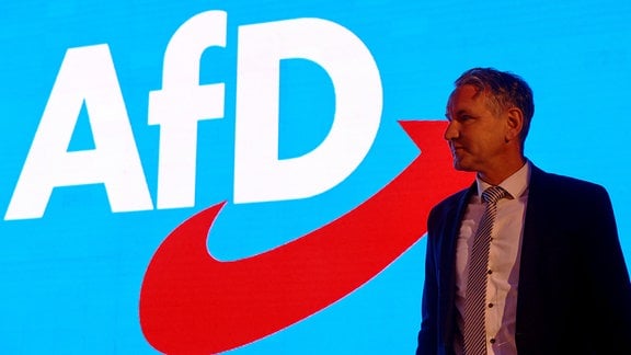 Björn Höcke steht vor einem großen blauen Hintergrund mit dem AfD-Logo.