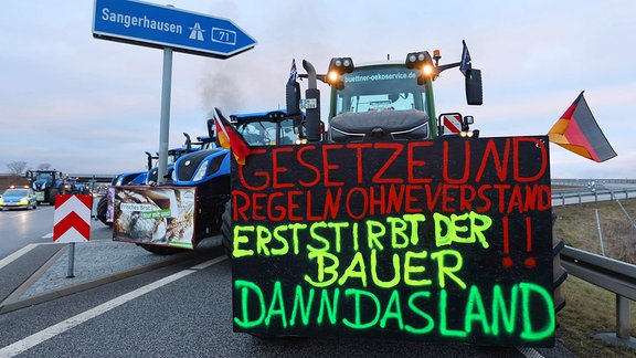 Traktoren stehen in der Auffahrt Kölleda in Richtung Sangerhausen zur Autobahn A71  - an der Front eines Traktors ist zu lesen: "Gesetze und Regeln ohne Verstand, erst stirbt der Bauer, dann das Land"