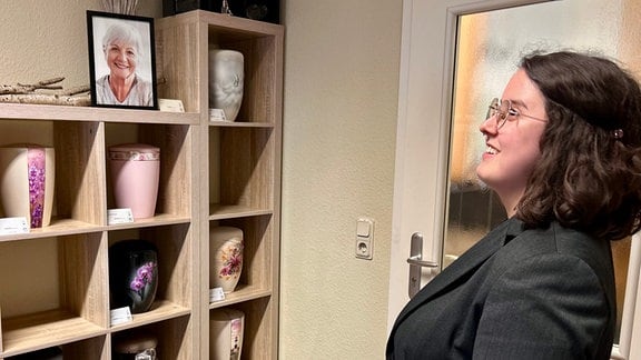 Bestatter-Azubi Franziska Ritscher steht in einem hellen Raum vor einem Regal mit Urnen. Sie schaut seitlich in das Regal und lächelt dabei.