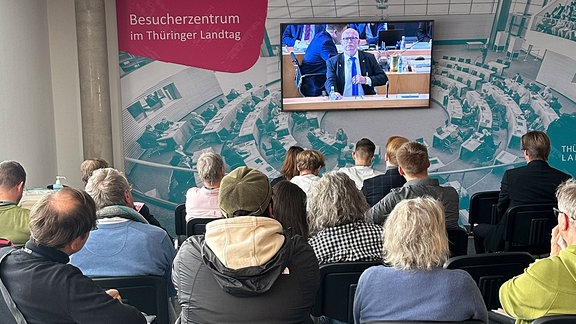  Menschen sitzen vor einem großen Fernseher im Besucherzentrum des Thüringer Landtags