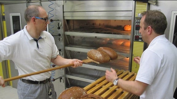 Zwei Bäcker in weißen Oberteilen und schwarz-weiß karierten Hosen stehen vor einem Industriebackofen in der JVA Tonna und heben frisch gebackene Brote heraus