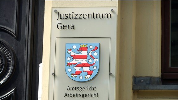 An einer gelb gestrichenen Hausfassade ist ein Schild angebracht mit der Aufschrift: Justizzentrum Gera, Amtsgericht, Landgericht. Außerdem ist das Thüringer Landeswappen auf dem Schild