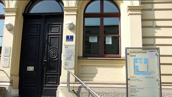 Eingangsbereich des Justizzentrums in Gera. Zu einem gelb gestrichenen Haus aus dem 19. Jahrhundert führt eine Treppe mit mehreren Stufen zu einer dunkelbraunen Holztür hinauf. Beiderseits der Tür sind mehrere Schilder an der Hauswand angebracht. Auf einem der Schilder steht: Justizzentrum Gera