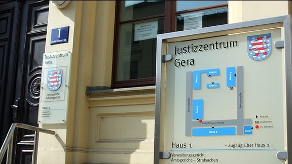 Vor einem gelb gestrichenen Haus steht ein großes Schild mit der Aufschrift Justizzentrum Gera. Unter diesem Schriftzug befindet sich ein Lageplan des Gebäudekomplexes