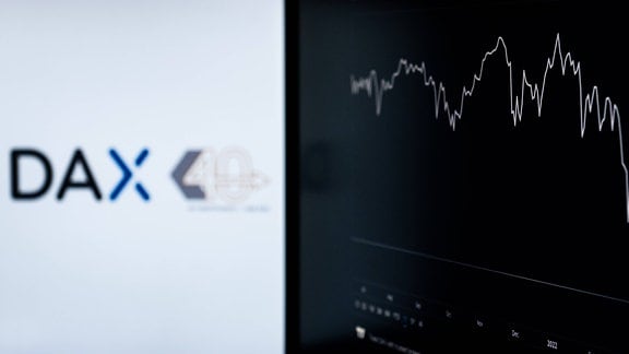 Das Logo des Deutschen Aktienindex DAX ist neben einem Linienchart des DAX auf Bildschirmen in einem Büro zu sehen.