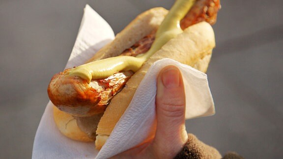 Eine Hand hält eine Bratwurst mit Senf in einem Brötchen