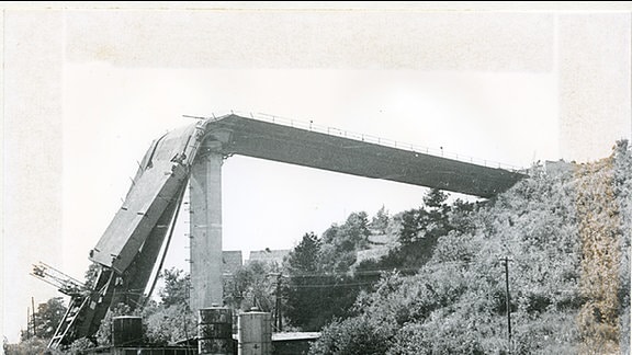 Schwarz-weiß-aufnahme eine Straßenbrücke über einen Grund, die nach dem ersten Pfeiler nach unten abgeknickt ist.