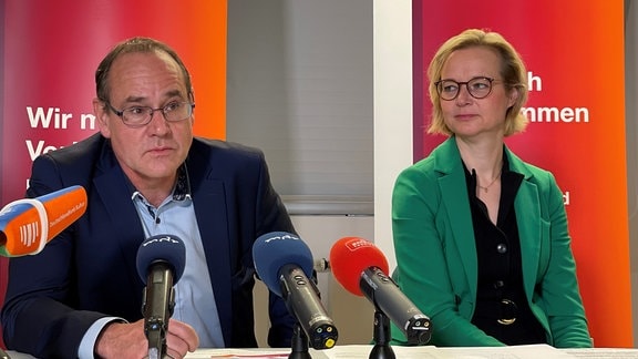 Ein Mann und eine Frau sitzen bei einer Pressekonferenz an einem Tisch. Vor ihnen stehen vier Mikrofone.