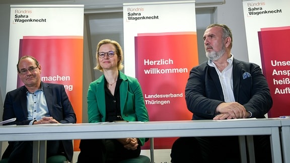 Zwei Männer und eine Frau sitzen bei einer Pressekonferenz an einem Tisch. 