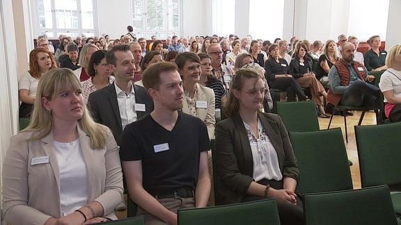 Menschn sitzen in Reihen hintereinander und tragen Namensschilder.