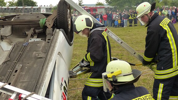 Drei Feuerwehrmänner stehen vor einem auf dem Dach liegenden weißen Pkw. Sie schicken sich an, mit einem Schneidgerät das Auto zu öffnen.