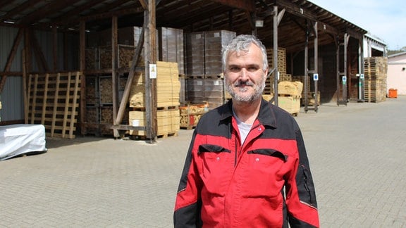 Ein Mann in rot-schwarzer Arbeitskleidung auf dem Hof der Holzwerkstatt.