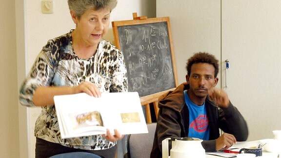 Die Deutschlehrerin Regina Claving hält bei einer Unterrichtsstunde in Deutsch für Flüchtlinge ein geöffnetes Buch, so dass ihre Zuhörer hineinsehen können; im Hintergrund sind ein Mann mit dunkler Hautfarbe und eine Tafel zu erkennen