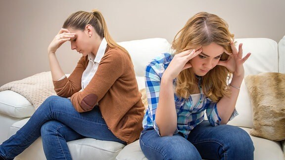 Zwei Frauen sitzen auf einem Sofa mit dem Rücken zueinander und schauen verärgert.