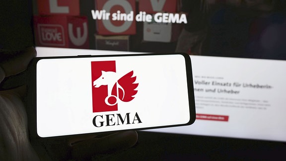 Person hält Smartphone mit Logo der deutschen Verwertungsgesellschaft GEMA vor Website. Fokus auf Telefondisplay.