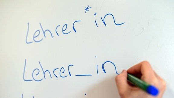 Eine Hand schreibt "Lehrer_in" auf eine Tafel.