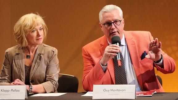 Angela Klepp-Pallas, eine Frau mit tittellangen blonden Haaren und Guy Montavon, ein Mann on orangenem Anzug mit Brille