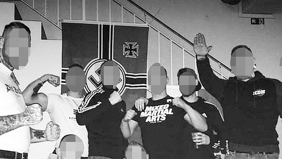 Mehrere junge Männer stehen vor einer Fahne mit Hakenkreuz. Einer reckt den Arm zum Hitlergruß.