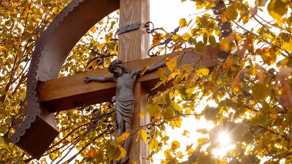 Jesus Christus Figur an einem hölzernen Kreuz wird von einem Sonnenschein durch bunte Herbstblätter angestrahlt
