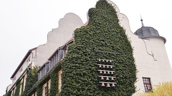 An einer Hausfassade hängt ein Glockenspiel inmitten hochkletternder Pflanzen.