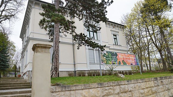 Eine Treppe führt zu einer Villa. Rechts und links der Villa stehen alte Bäume. In der Villa ist das Kunshaus Apolda untergebracht. Ein große Plakat macht auf die Ausstellung Hermann Hesses aufmerksam. 