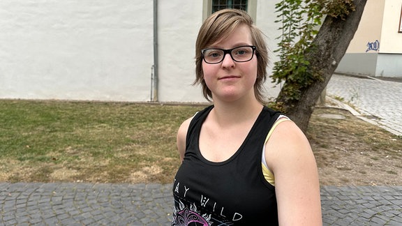 Die junge Frau, Lena Mehlgarten lächelt. Sie hat kurze blonde Haare und trägt eine Brille mit schwarzem Gestell und ein schwarzes Top mit bunten Aufdruck. 