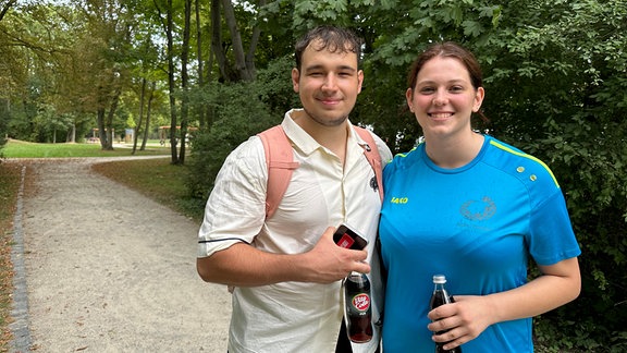 In einem grünen Park steht ein junges Paar, das in die Kamera lächelt. Rechts steht Jacob Hanika. Er trägt ein gelbes Kurzarmhemd, hat dunkle Haare und trägt einen Rucksack. Dich neben ihm steht seine Freundin Lenka Loszkorih in einem blauen Sportshirt.Beide halten eine Vita-Cola in der Hand.