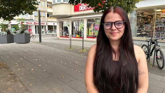 Sophie Schröder lächelt in die Kamera. Die junge Frau trägt eine schwarz umrandete Brille und sie hat lange dunkle Haare. Im Hintrergrund sieht man Geschäfte der Innenstadt von Sömmerda.