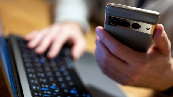 Ein Mann sitzt am Laptop und hält ein Smartphone mit in der Hand.