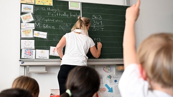 Schüler einer Grundschule arbeiten in einem Klassenzimmer an Mathematikaufgaben. Die Lehrerin schreib an der Wandtafel.