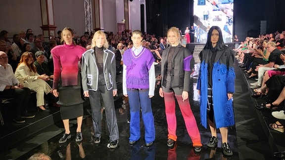 Fünf junge Frauen auf einem schwarzen Laufsteg präsentieren eine Modekollektion.