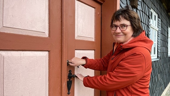 Eine Frau in einer roten Jacke steht an der Eingangstür eines alten Fachwerkhauses. 