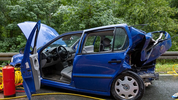 Ein blaues Auto steht nach einem Unfall zerstört auf einer Straße.