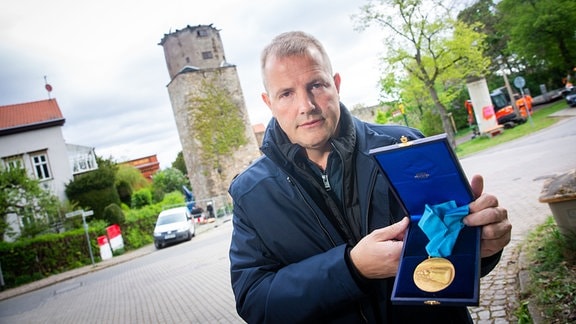 Ein Mann steht vor einem Turm und hält eine Schachtel mit einer Goldmedaille in die Kamera.