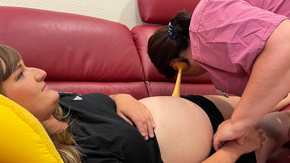 Eine Hebamme horcht am Bauch einer schwangeren Frau