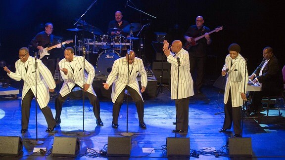 Fünf Sänger in hellen Jackets stehen auf einer Bühne und singen. Im Hintergrund spielen mehrere Musiker
