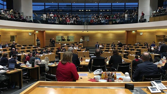 Im Plenarsaal des Thüringer Landtags sitzen mehrere Menschen an Tischen und im Zuschauerraum