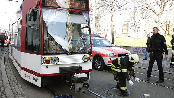 Eine Straßenbahn mit beschädigter Frontscheibe steht neben einem Notarzt-Wagen in einer Straße, auf der sich ein Feuerwehrmann zu Papierstücken hinunterbeugt