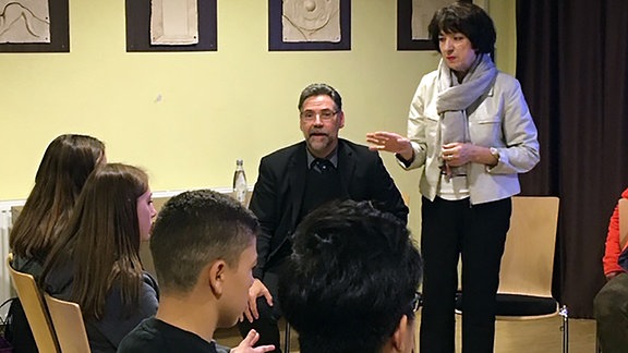 Eine Frau mit schwarzen, halblangen Haaren und einer weißen Jacke steht neben einem Mann und redet, vor ihr sitzen Jugendliche in einem Publikum