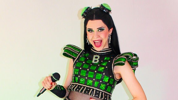 Eine junge Frau in einem zweiteiligen grün-glitzerndem Kostüm. Sie hält ein Mikrofon in der rechten Hand und stellt die linke Hand in die Hüfte. Sie wirkt euphorisch. 