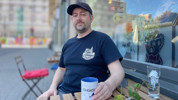 Ein Man mit Bart, schwarzem T-Shirt und schwarzem Baseballcap sitzt an einem Tisch vor einem Ladenfenster. In der Hand hält er eine Kaffeetasse.