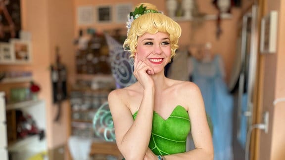 Eine junge Frau im Kostüm von "Tinkerbell" aus dem Disneyfilm "Peter Pan". Sie trägt eine blonde Perücke, Feenflügel und ein grünes Kleid. 