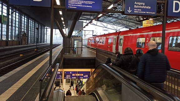 Menschen auf einer Rolltreppe neben einem Zug im Bahnhof.