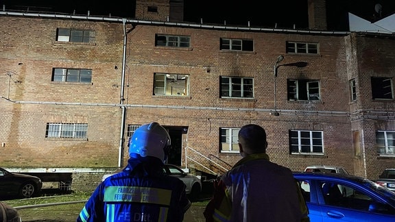 Zwei Feuerwehrleute blicken nachts auf ein verrustes und ausgebranntes Ziegelsteinhaus