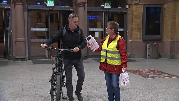 DGB-Mitarbeiterin reicht einem Mann mit Fahrrad am Hauptbahnhof Erfurt eine Tüte.